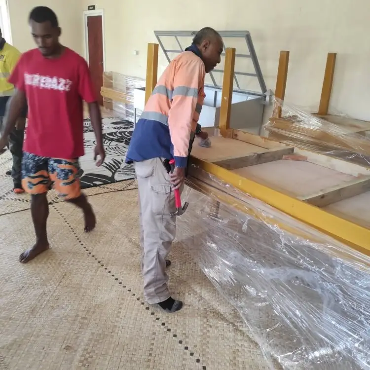 Senior PWD worker examines the table frame for any defects. A villager walks behind him.